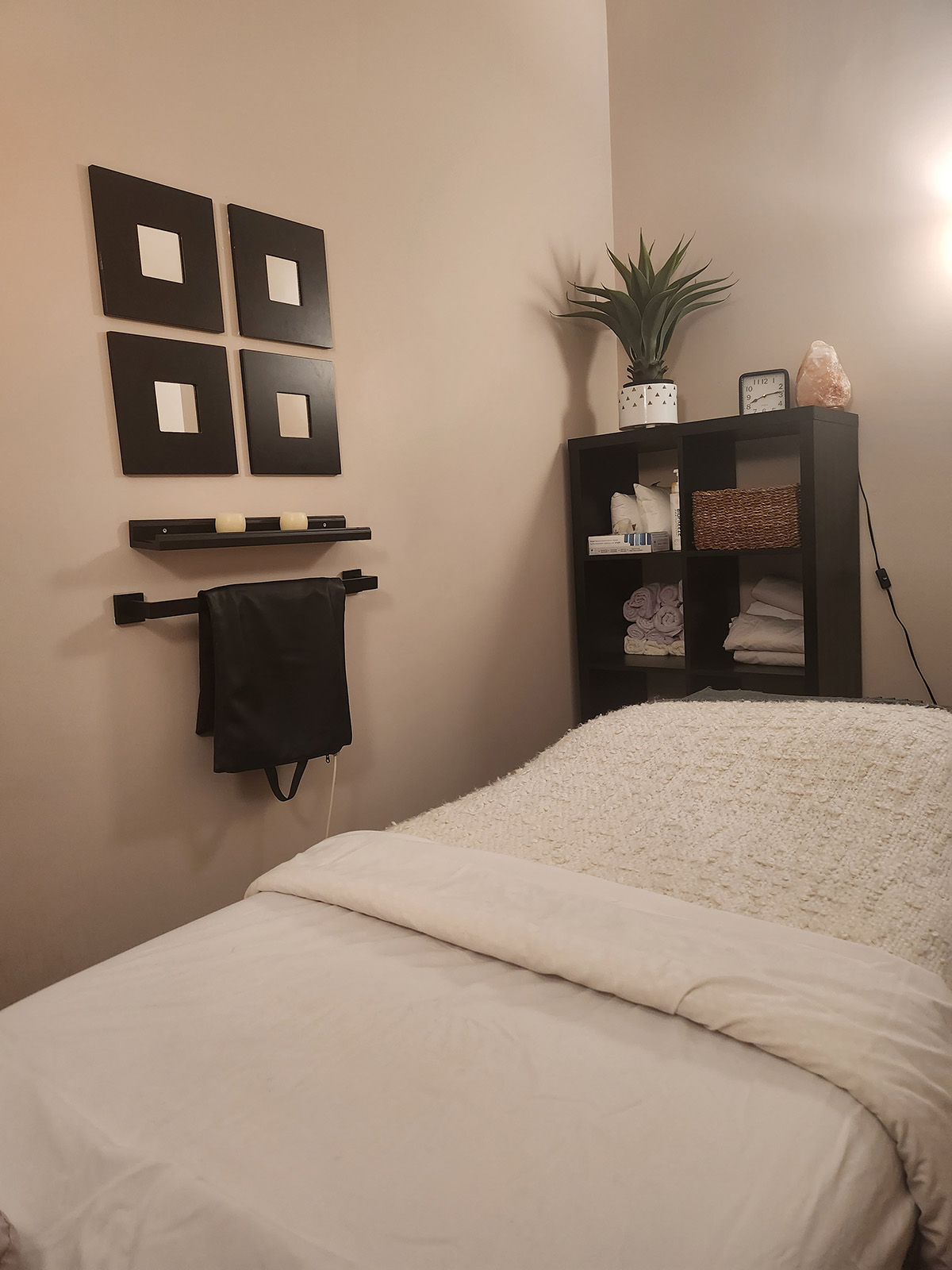 A cozy massage treatment room scene featuring a neatly made bed with white linens, a bookshelf with various items, a massage therapy salt lamp, and minimalist black square wall decor.